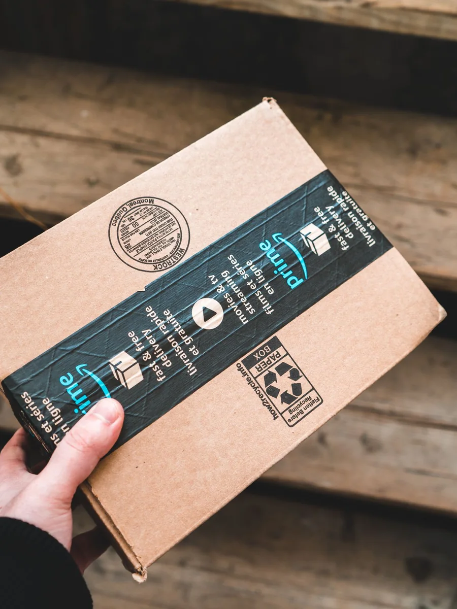 A gloved hand holding an Amazon Prime package above wooden steps at a delivery stop
